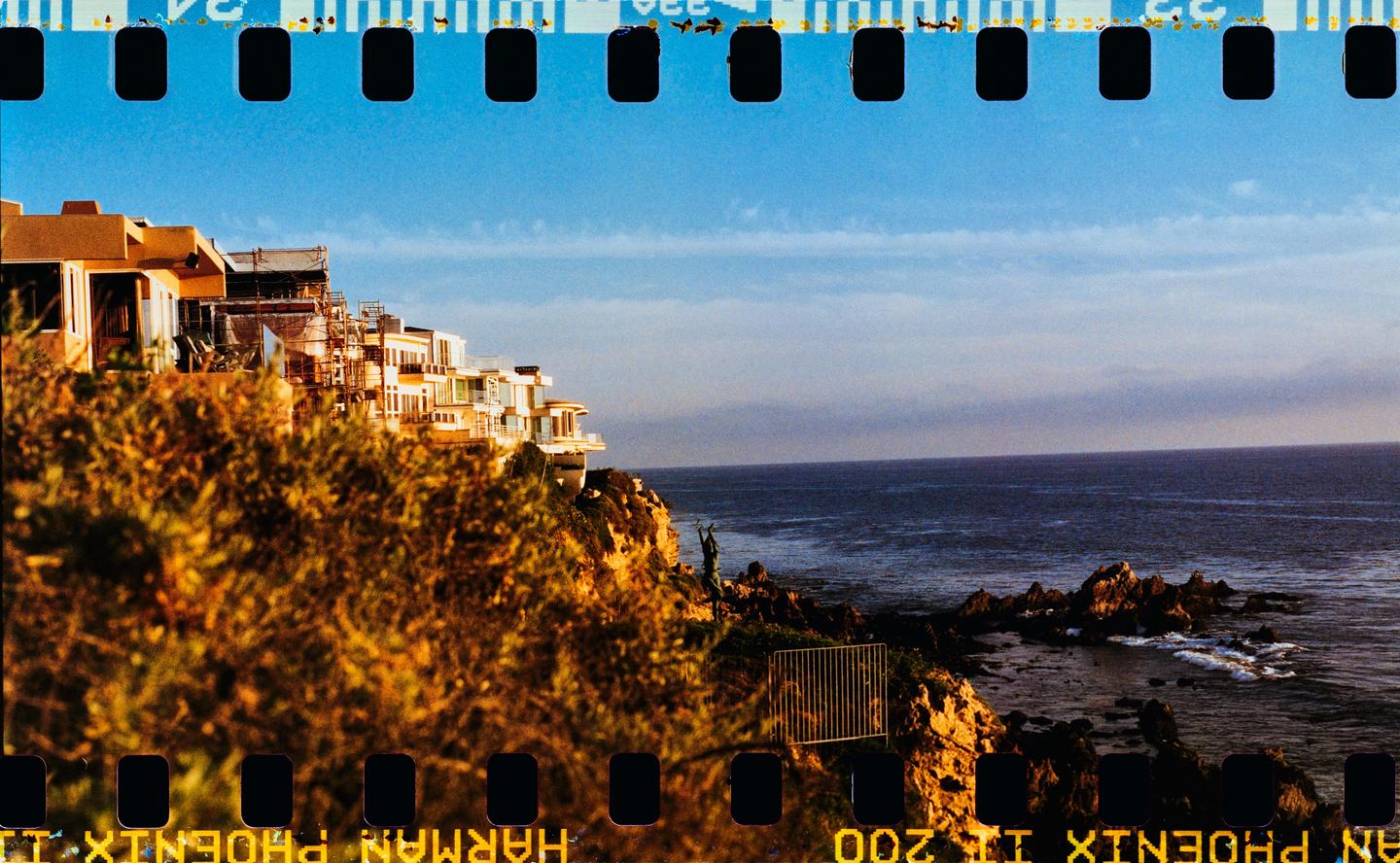 A row of houses sits on a cliff overlooking the ocean under a bright blue sky.