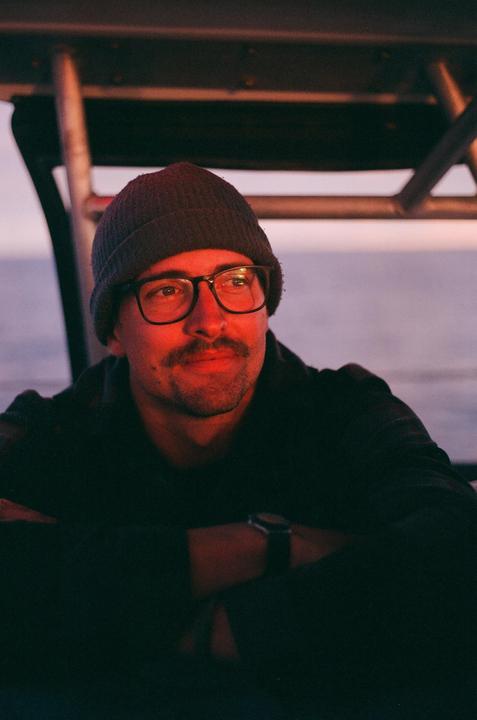 A man wearing a beanie and glasses looks to the right while sitting on a boat at sunset.