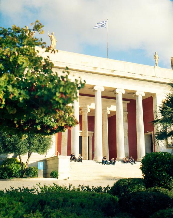 A group of people sit on the steps of a large building with columns and a Greek flag flying from the roof.