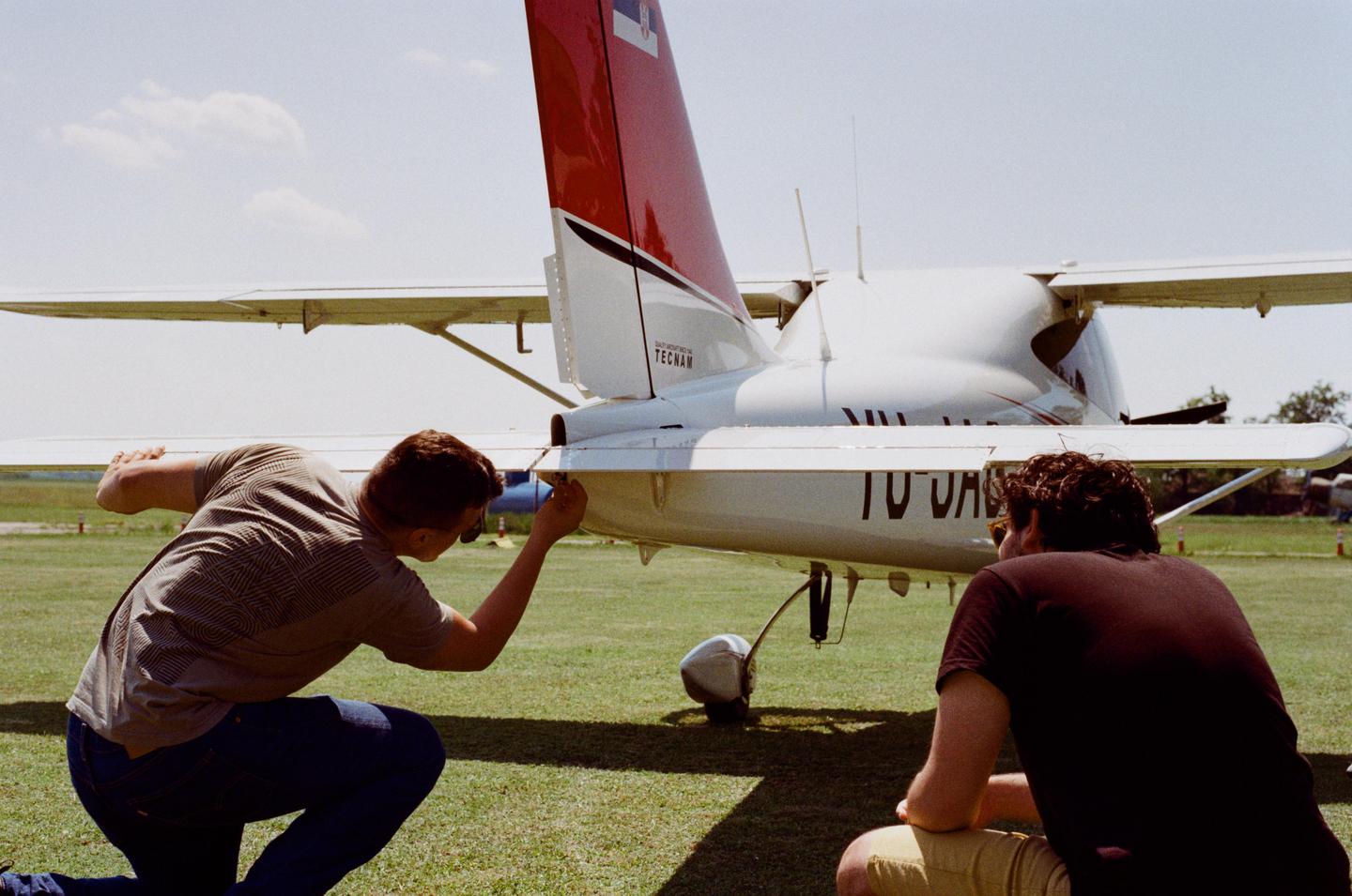 Two men are performing a pre-flight check on a small white airplane with a red tail.