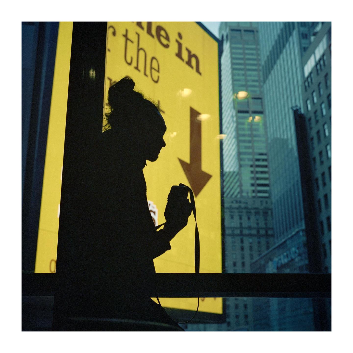 A person is silhouetted against a bright yellow sign with an arrow pointing down, with city buildings visible in the background.