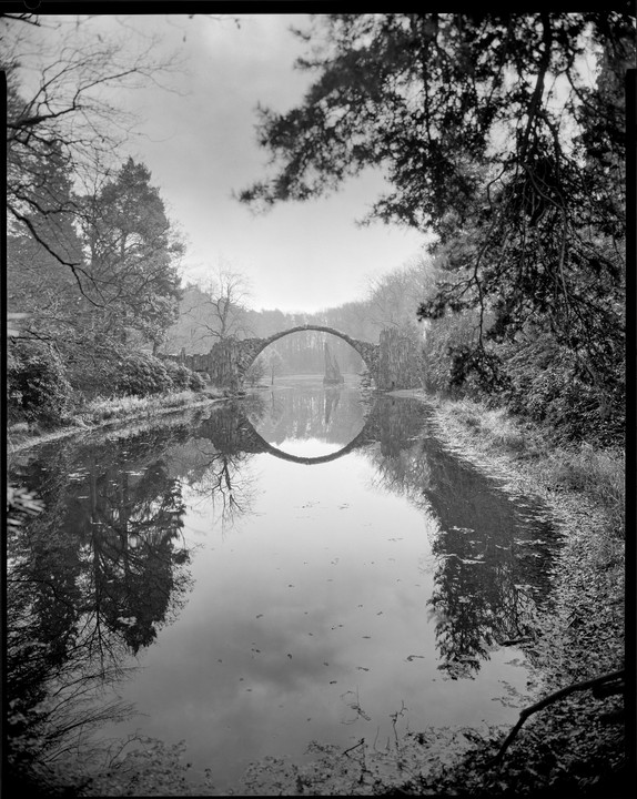 A circular stone bridge is reflected in the still water of a river, surrounded by trees on both banks.