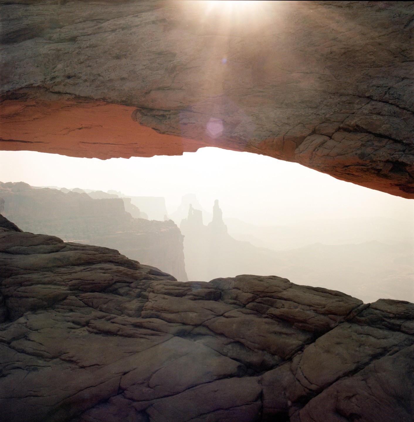 Sunlight shines through Mesa Arch, illuminating a hazy landscape with distant rock formations.