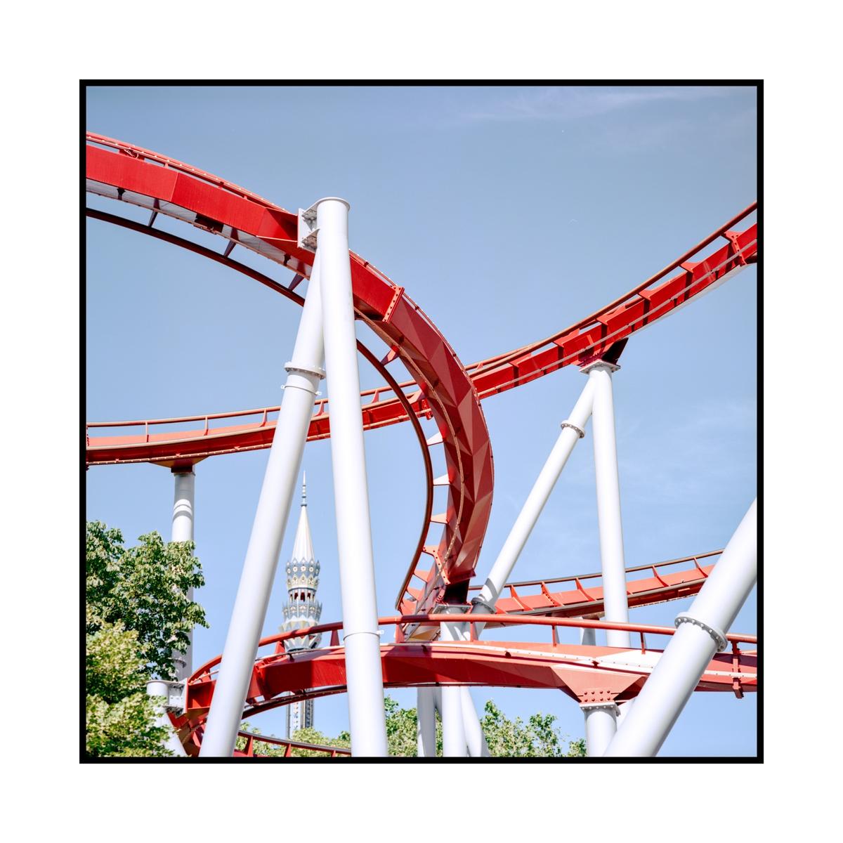 A red roller coaster track with white supports curves against a clear blue sky, with a white spire visible in the background.