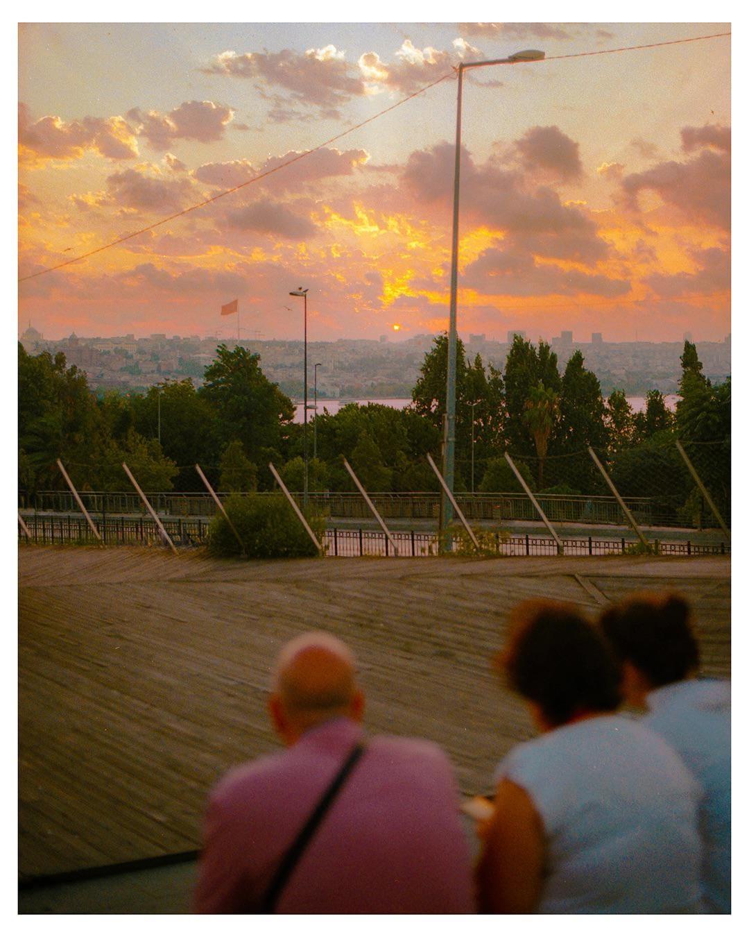 Three people sit on a wooden surface, looking out at a city skyline and water under a dramatic sunset sky.