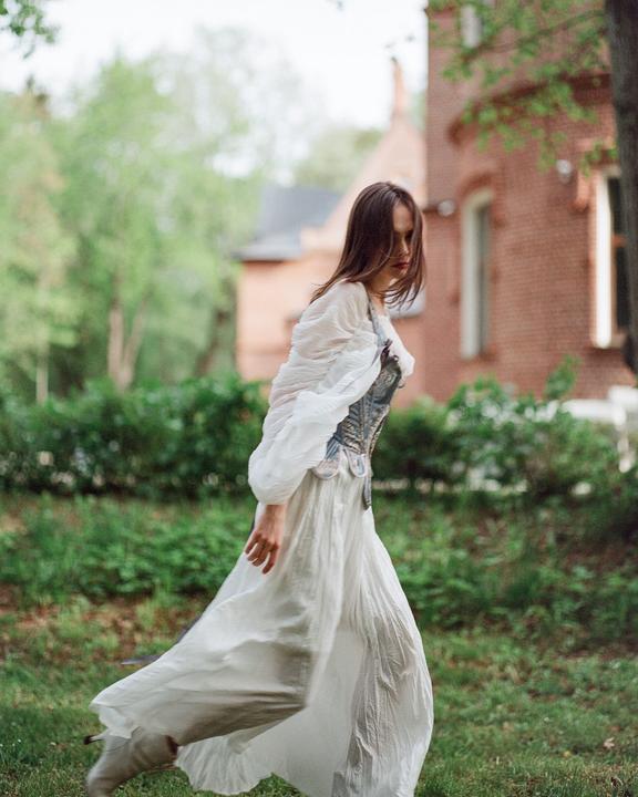 A woman in a white dress and corset walks through a grassy area with a brick building in the background.