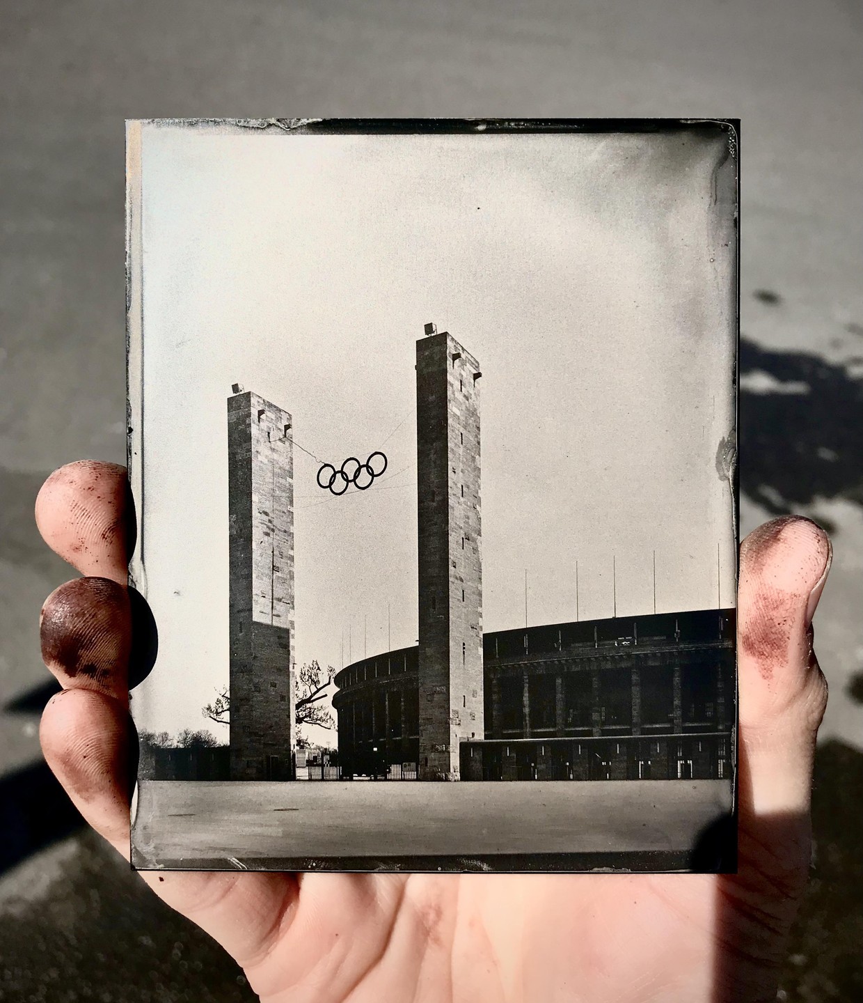 A hand holds a tintype of the Olympic Stadium in Berlin, with two tall towers and the Olympic rings visible.