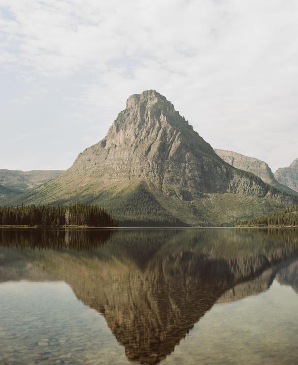 A large mountain is reflected in a still lake with a line of trees at its base.