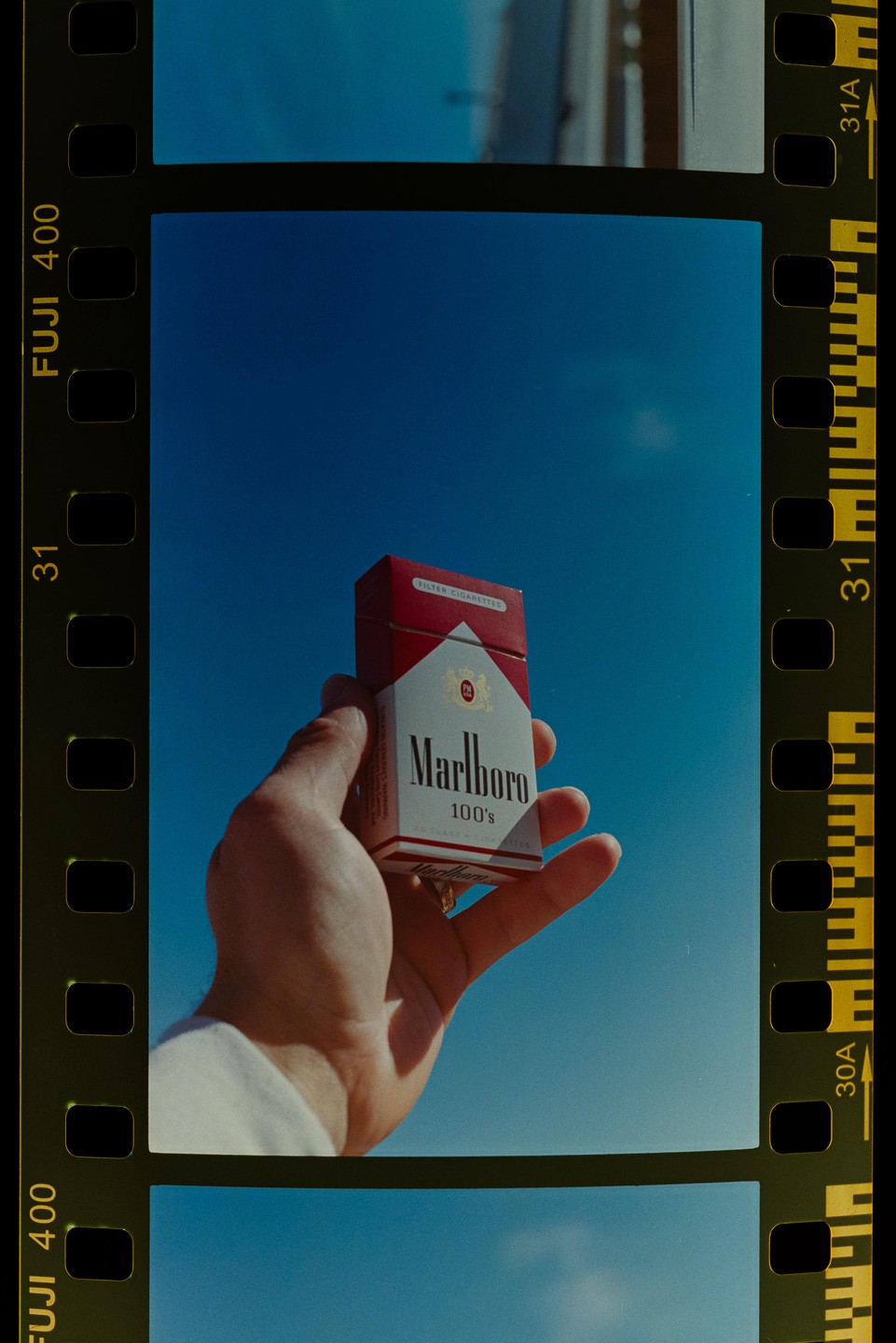A hand holds a pack of Marlboro cigarettes against a clear blue sky.
