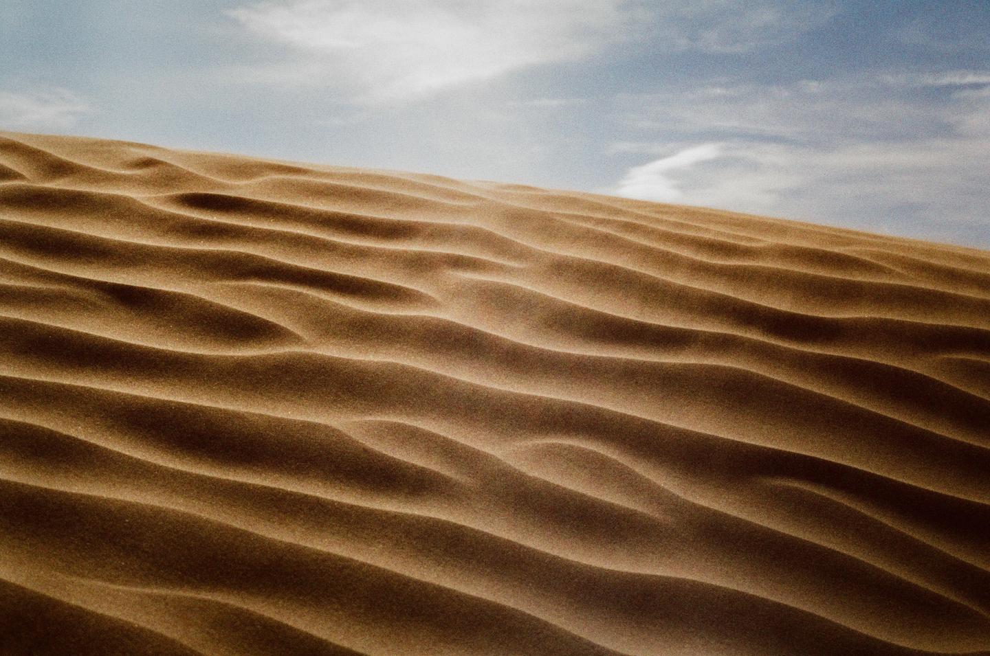A sand dune with ripples covers the lower two-thirds of the frame, with a blue sky and clouds above.