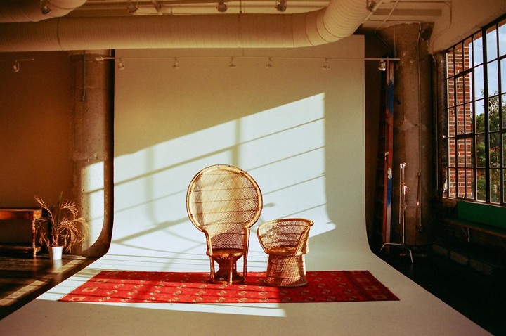 Two wicker chairs sit on a red rug in a sunlit studio with a large window showing a brick building outside.