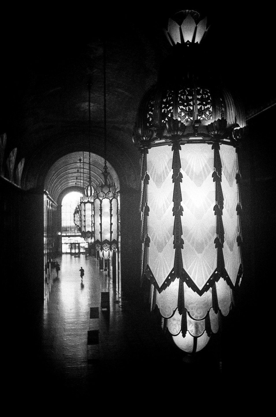 A large, ornate chandelier hangs in the foreground of a long, arched hallway with a person walking in the distance.
