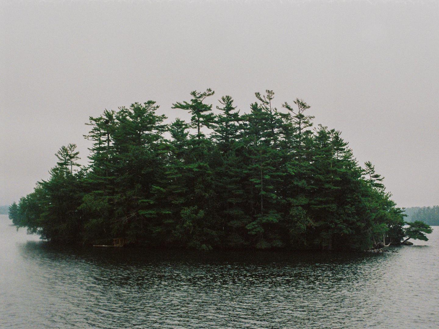 A small island covered in pine trees sits in the middle of a body of water under a hazy sky.