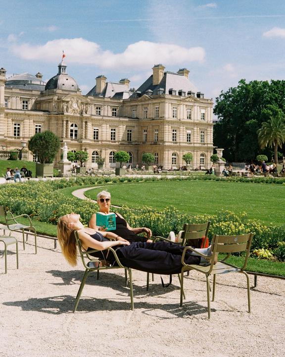 Two women relax in chairs on a gravel path in a park with a large building in the background.