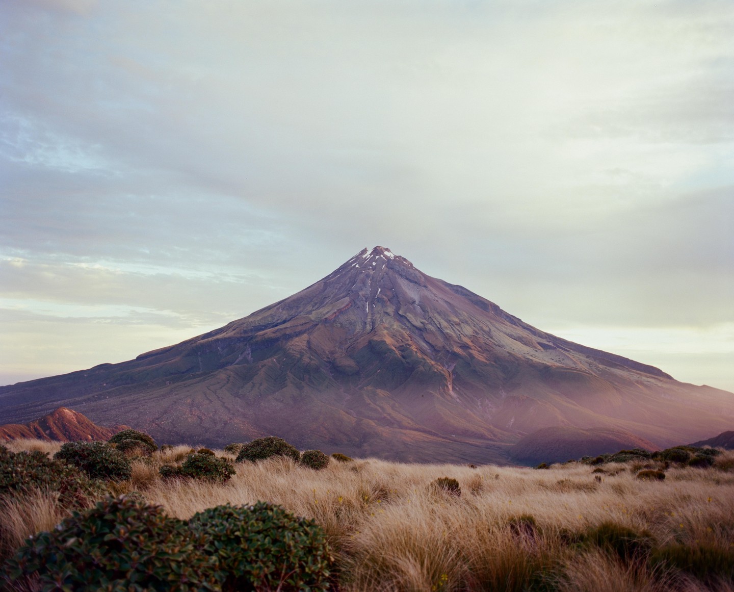A large mountain with a snow-capped peak rises above a field of dry grass and shrubs under a cloudy sky.