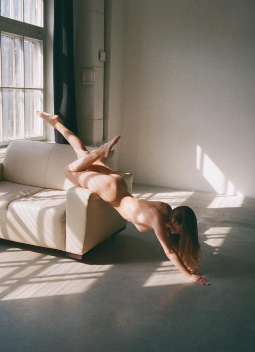 A nude woman is posed on a sofa with her legs in the air and her hands on the floor, with window light creating shadows on the wall and floor.
