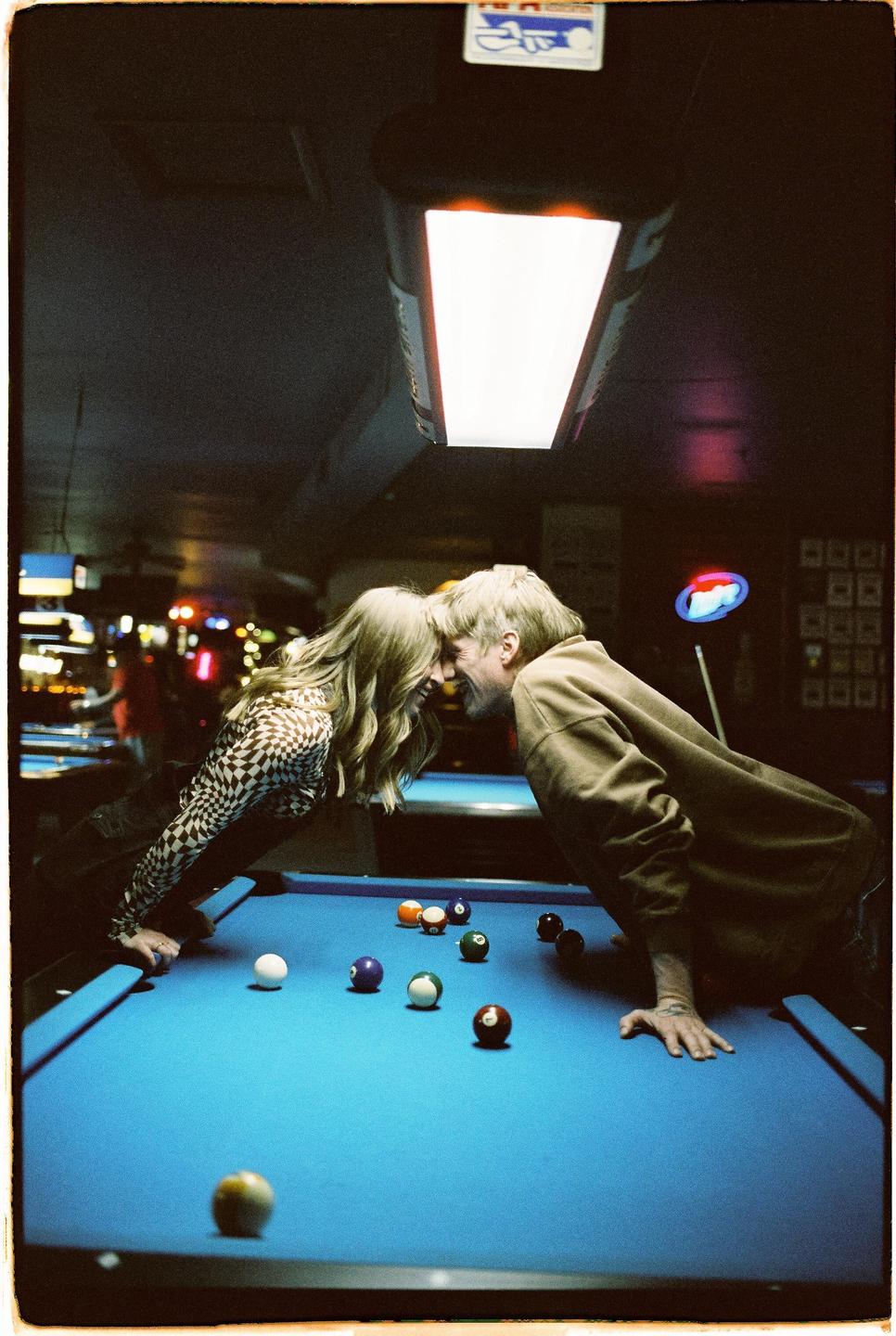 A couple leans over a pool table with their foreheads touching, surrounded by colorful pool balls.