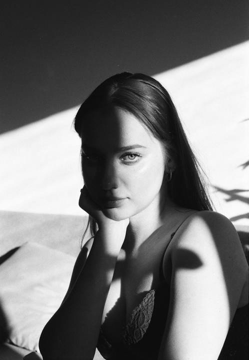 A woman with long hair rests her chin on her hand while sitting in a room with strong shadows.