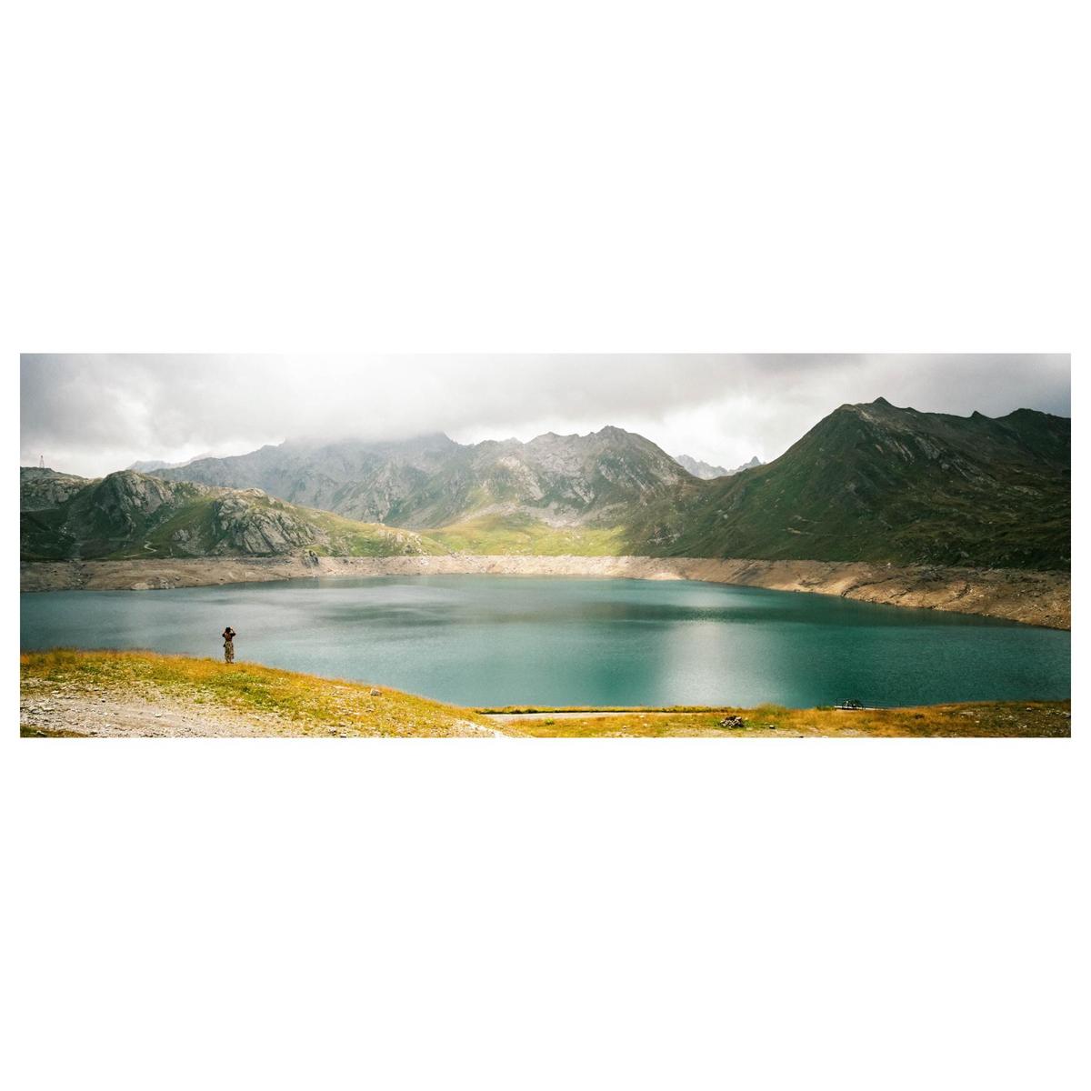 A person stands on a grassy hill overlooking a large lake surrounded by mountains under a cloudy sky.