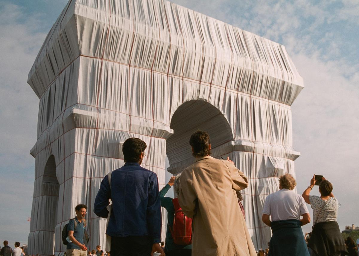 A crowd of people stands before a large, wrapped monument under a cloudy sky.