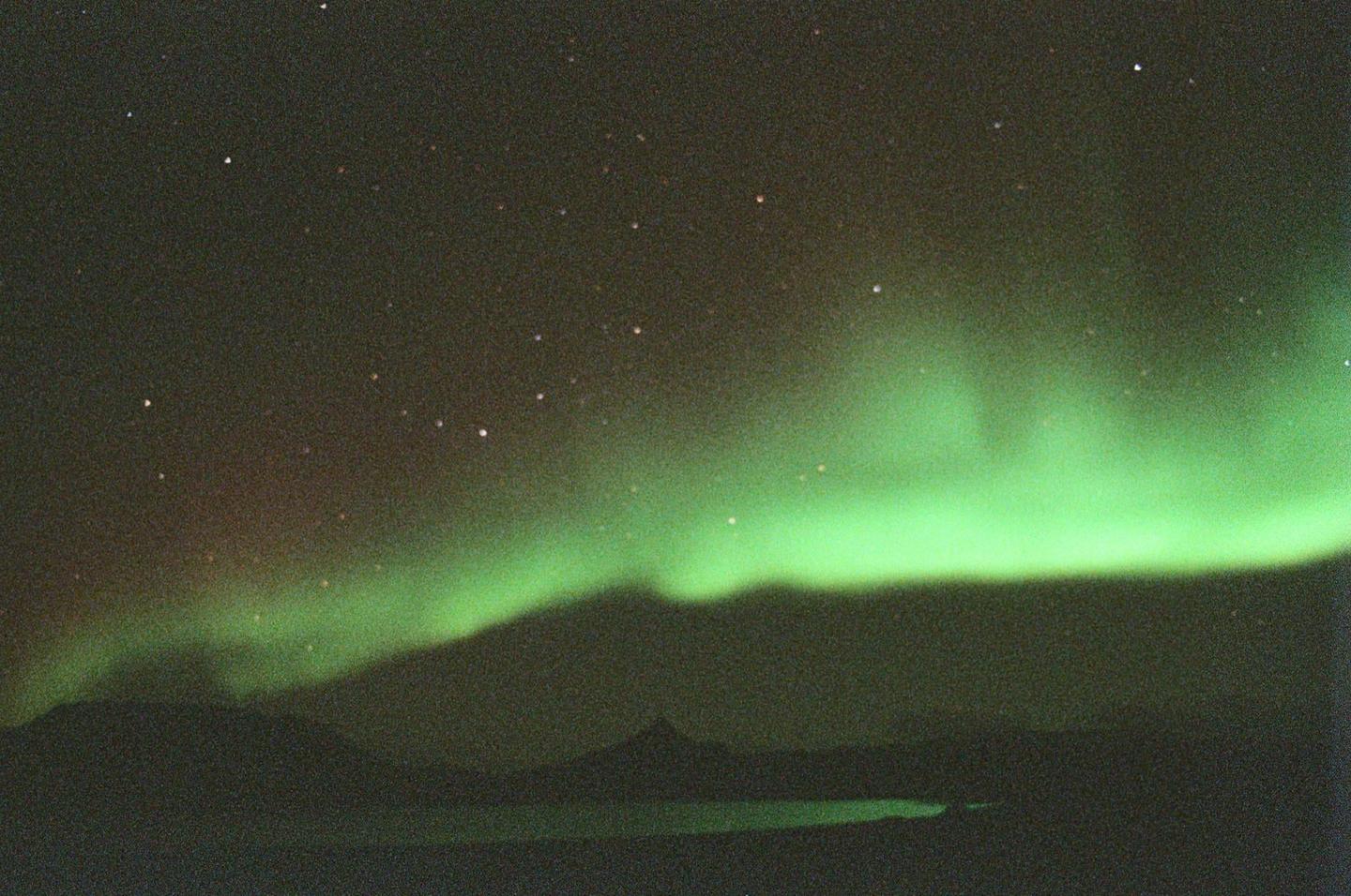 The aurora borealis glows green above a dark mountain range and a body of water under a starry night sky.