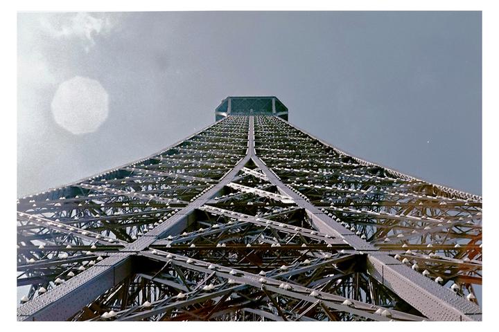 The Eiffel Tower is viewed from directly below, looking up at its intricate metal structure against a bright sky.