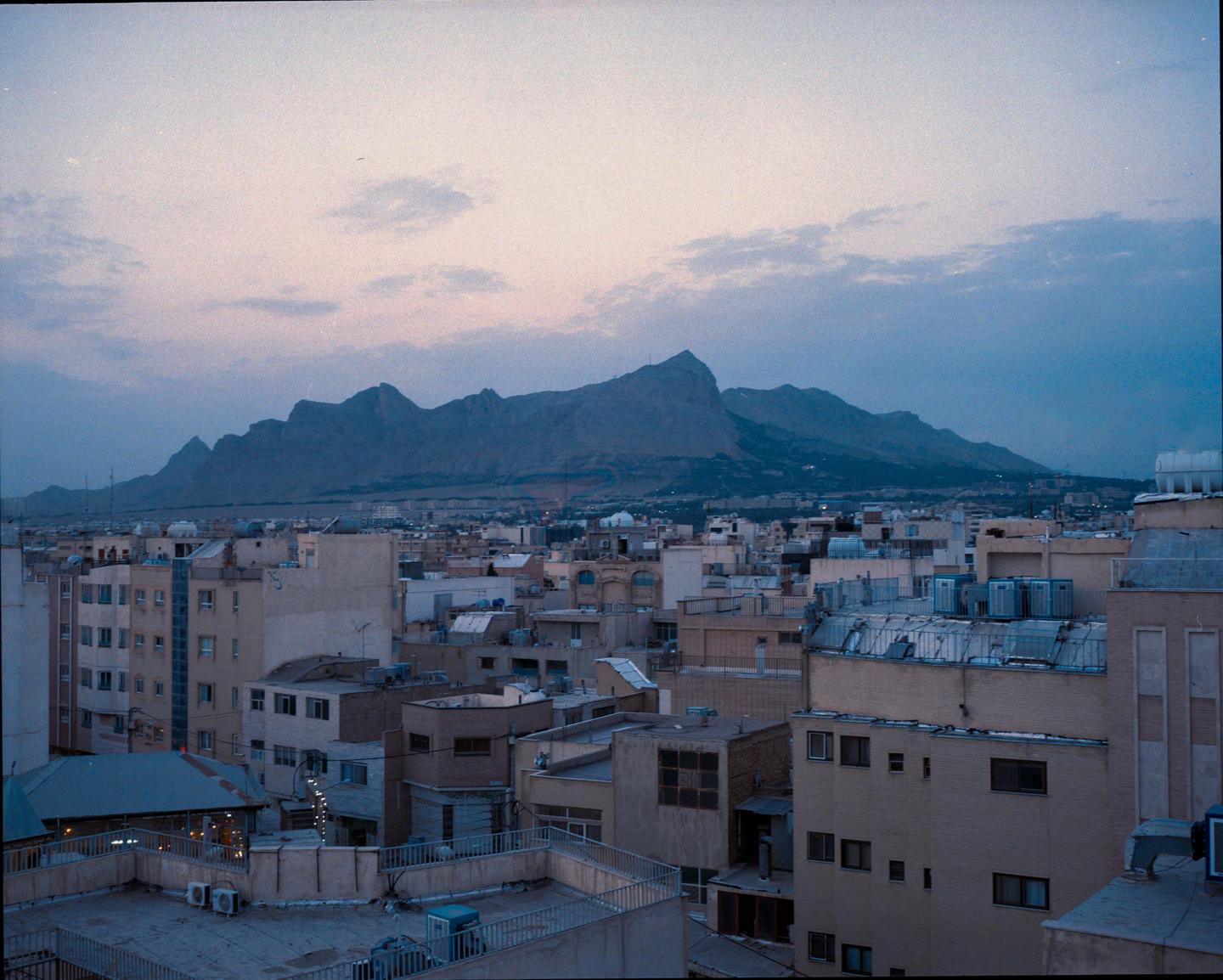 A city with many buildings and mountains in the background is visible under a cloudy sky.