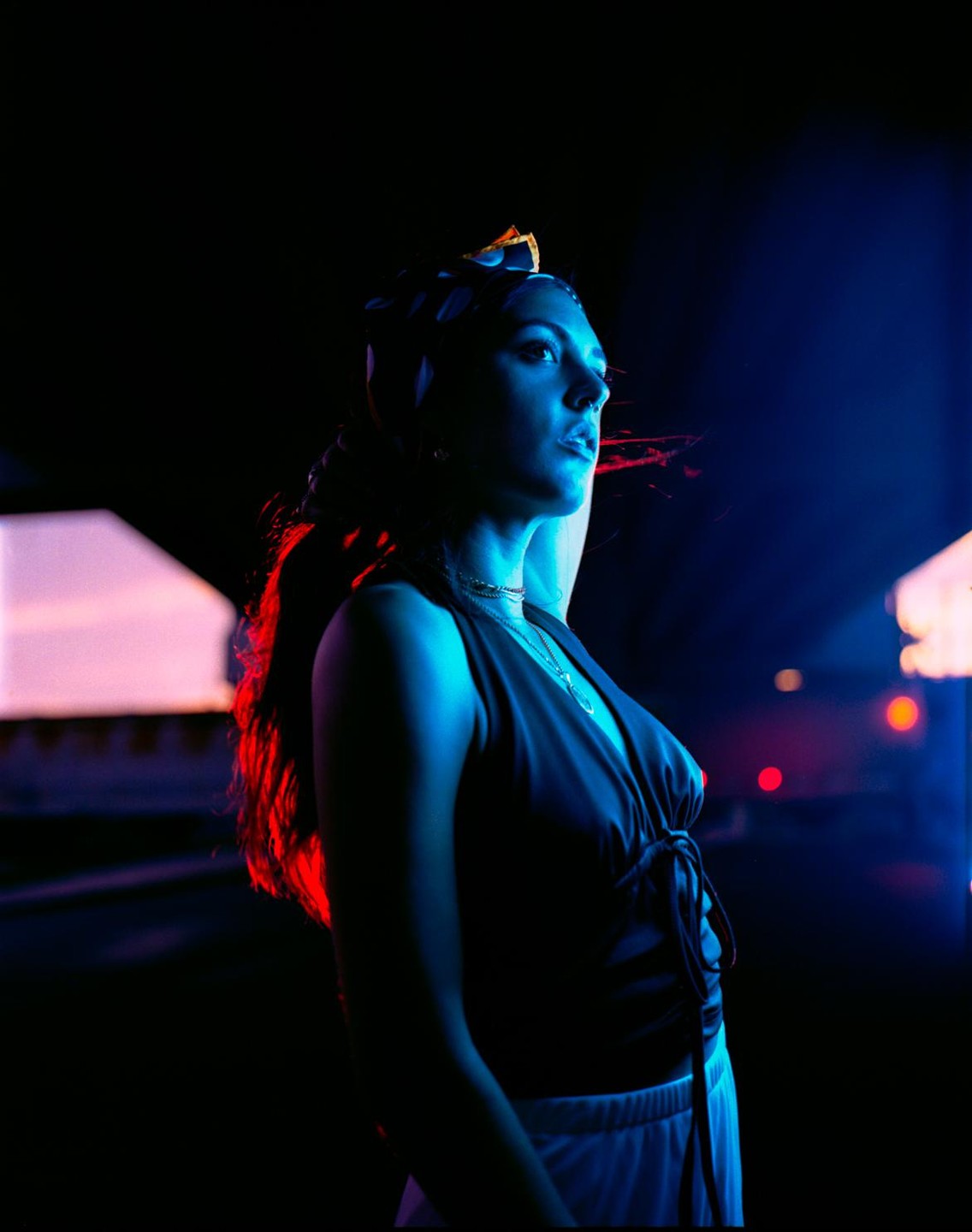 A woman with a headscarf and dark top stands in blue and red light at night.