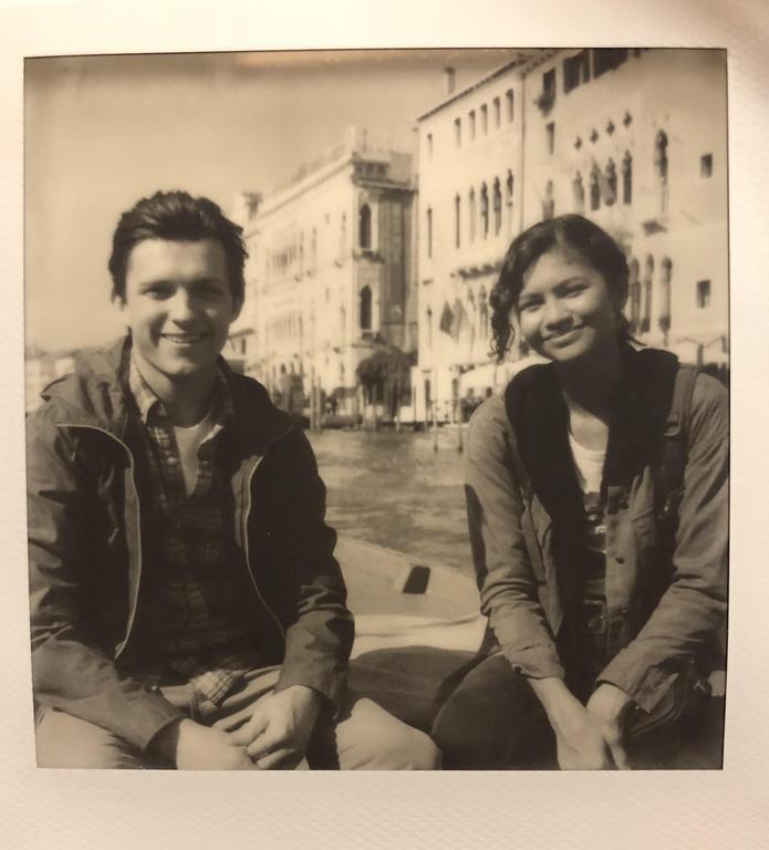 A man and a woman sit by a canal with buildings in the background.
