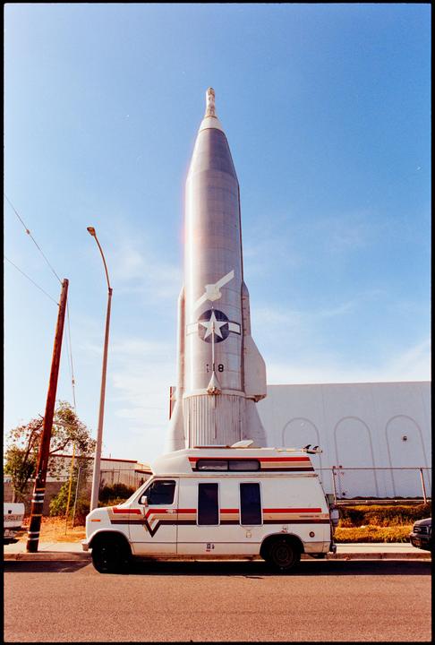 A white van with surfboards on its roof is parked in front of a large rocket under a clear sky.
