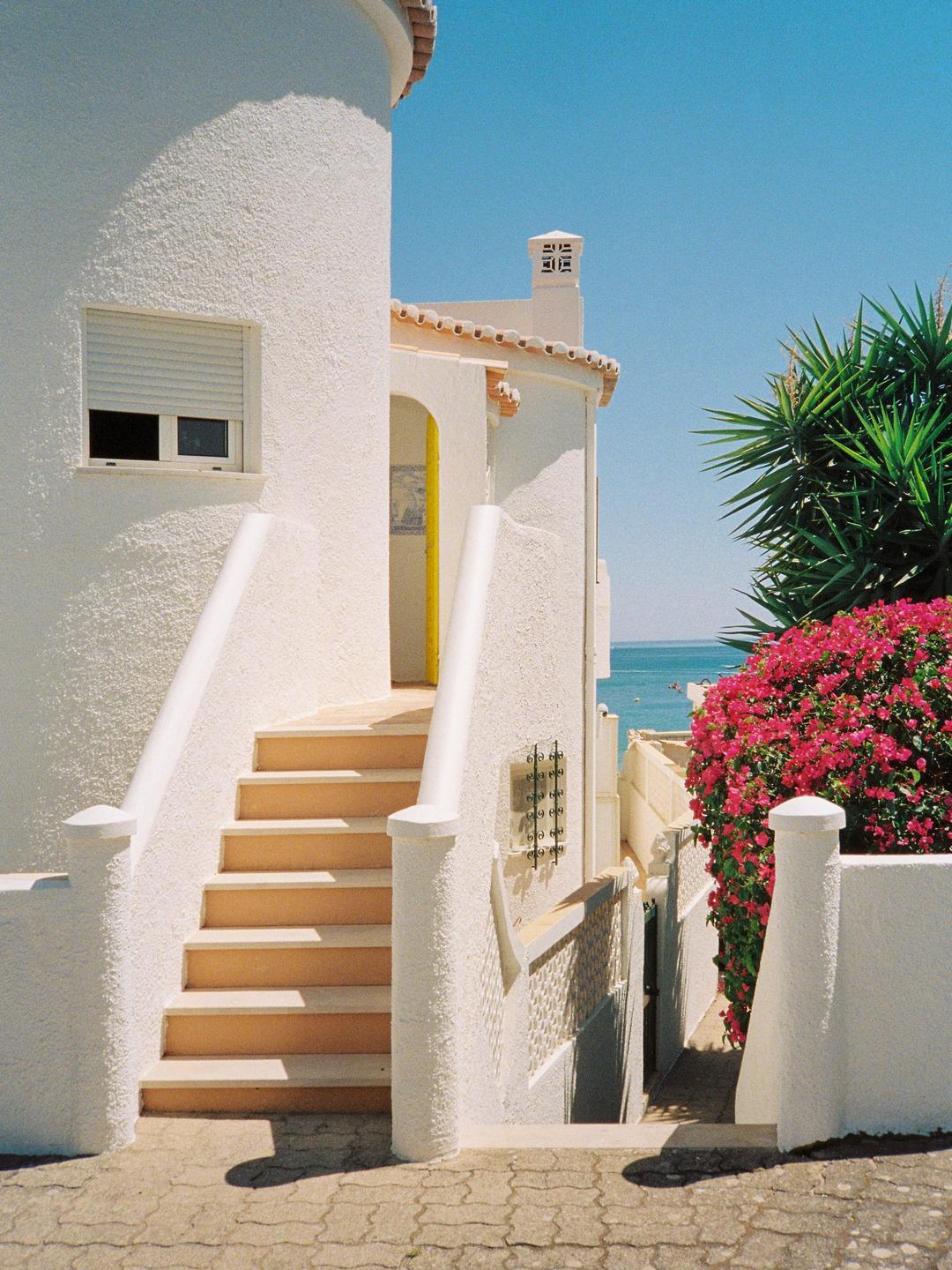 A white building with a yellow door and a staircase leads down to a narrow alleyway with the ocean visible in the distance.