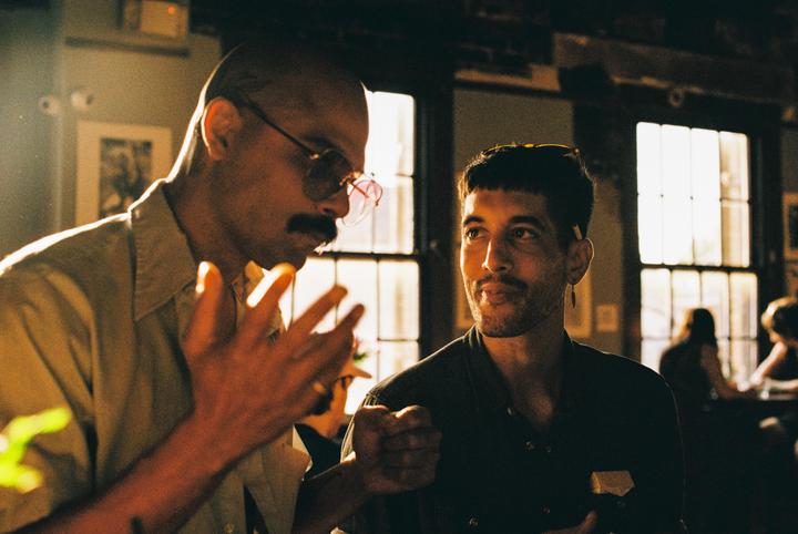 Two men are talking in a sunlit room with large windows in the background.