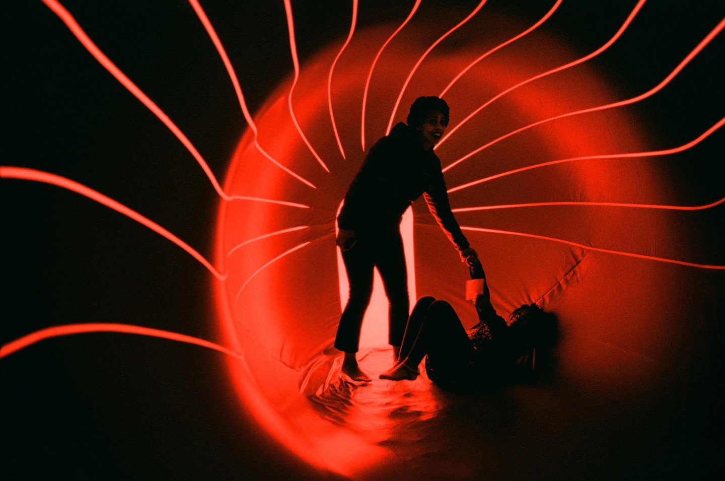 A woman helps another woman up from the ground inside a red-lit tunnel.