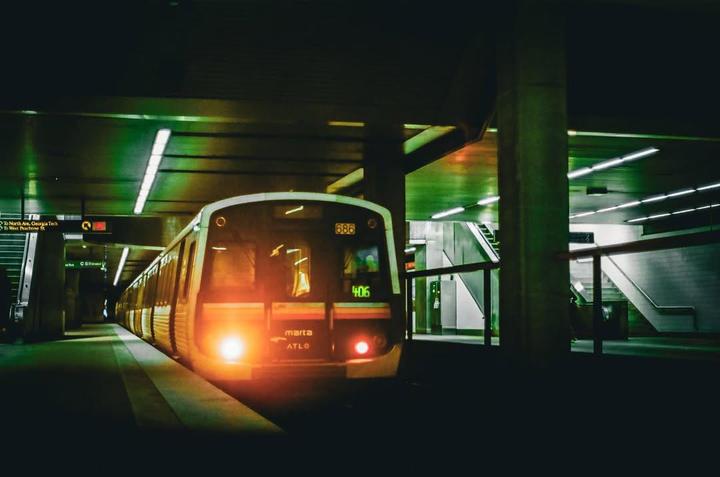 A train with bright headlights is stopped at a subway station platform with escalators and stairs visible in the background.
