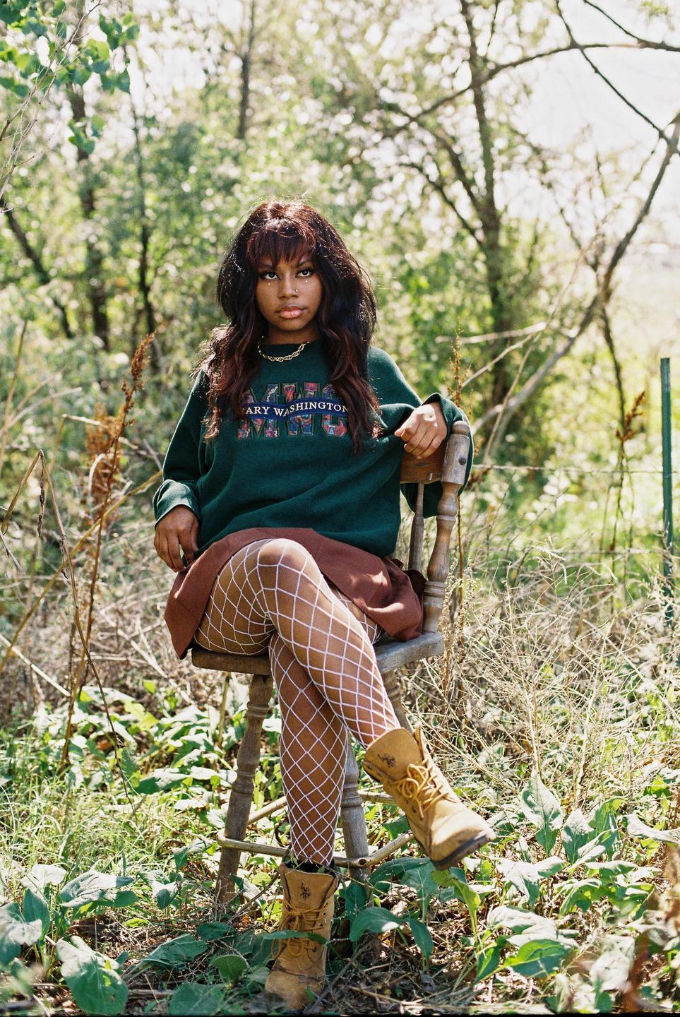 A woman wearing a green sweatshirt, brown skirt, white fishnet stockings, and tan boots sits on a wooden chair in a field.