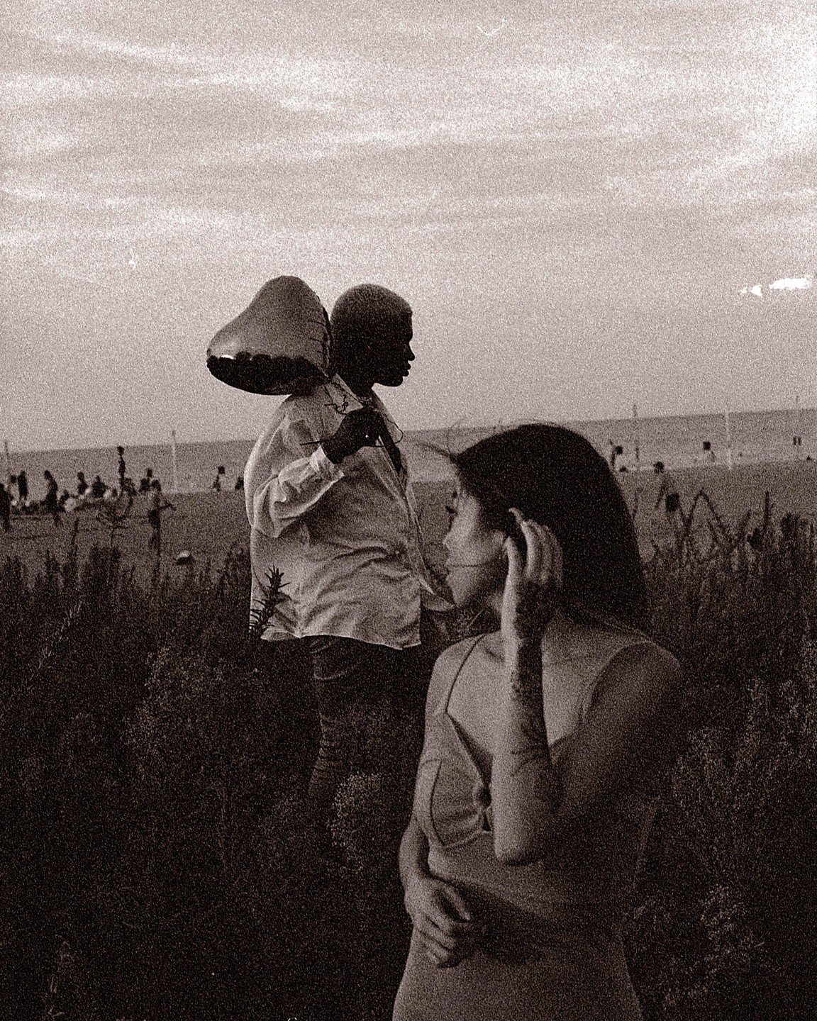 A woman stands in the foreground with a man carrying a heart-shaped balloon behind her, with a beach and ocean in the background.