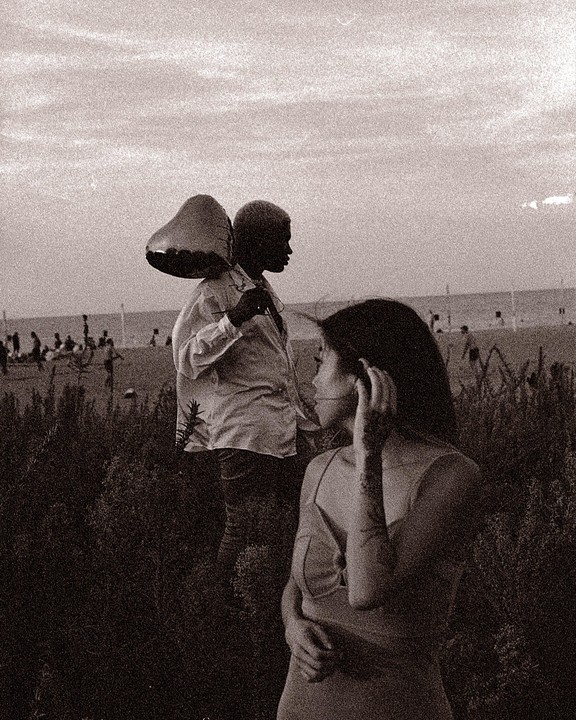 A woman stands in the foreground with a man carrying a heart-shaped balloon behind her, with a beach and ocean in the background.