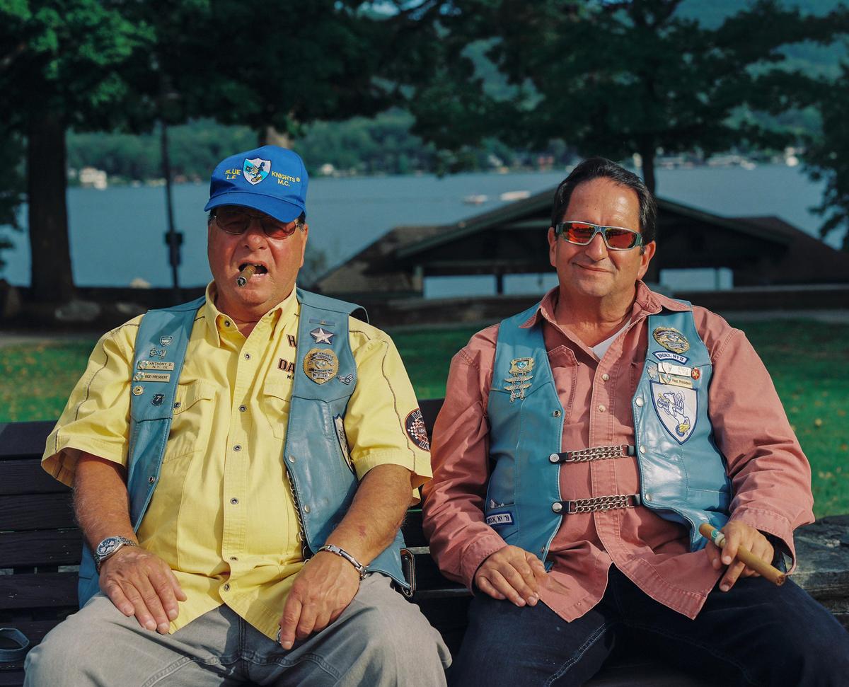 Two men wearing vests with patches are sitting on a bench with a lake and trees in the background.
