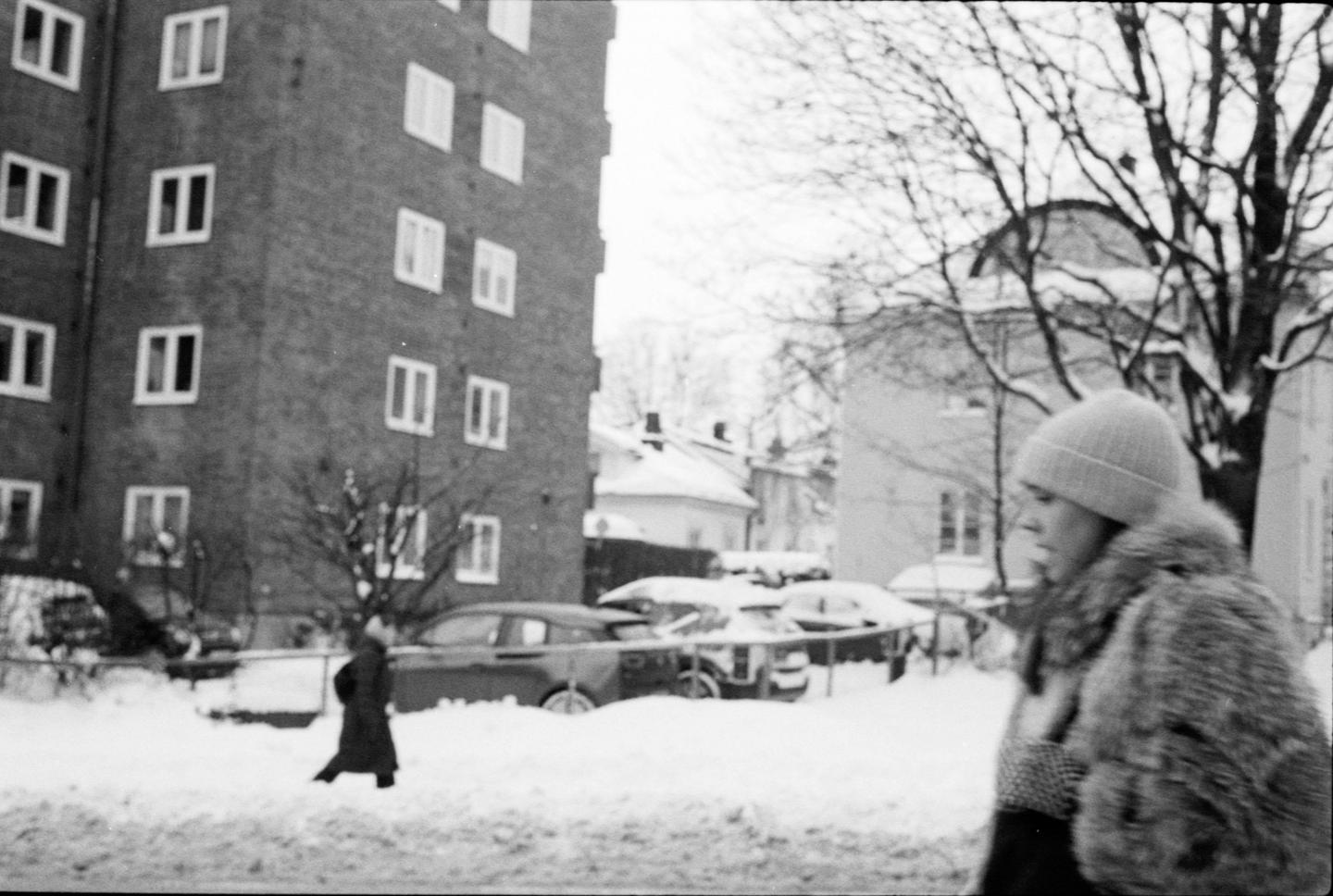 A woman in a fur coat and hat walks past a snow-covered street with buildings and cars.