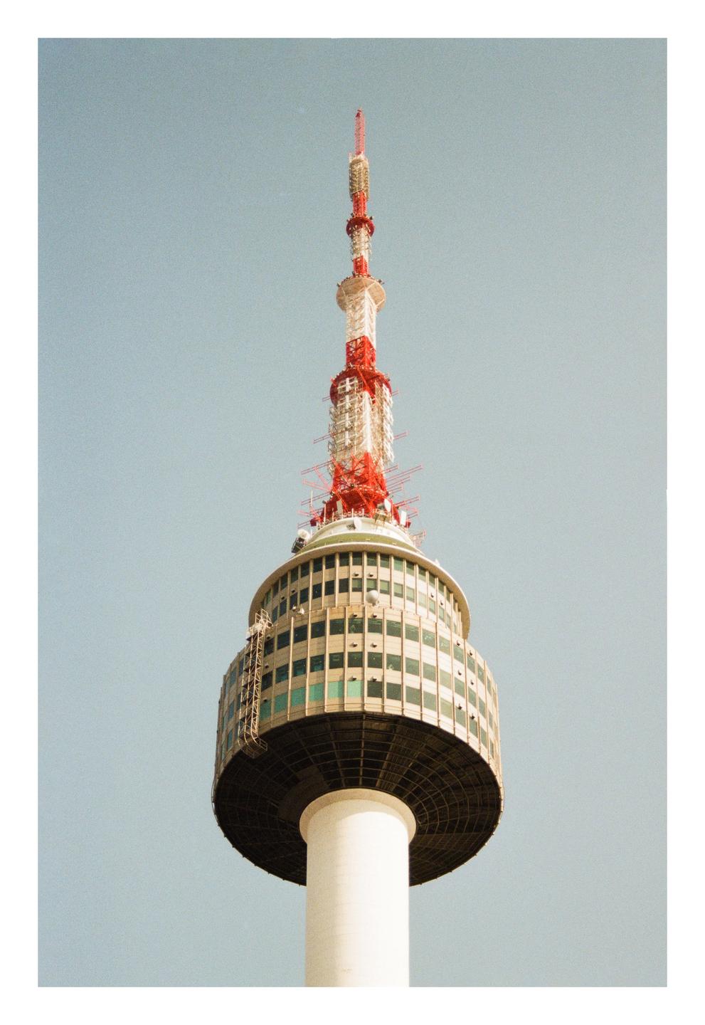 A tall white tower with a red and white antenna on top stands against a clear sky.