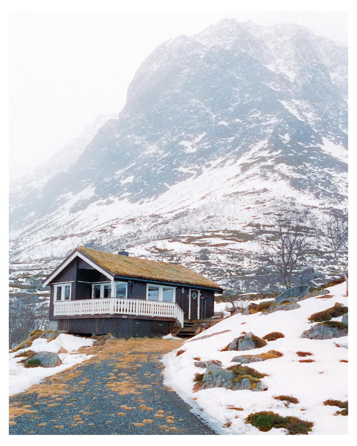 A small house with a grass roof sits on a snowy hillside with a large mountain in the background.