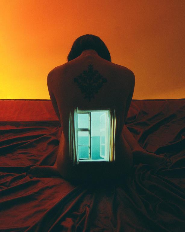A window showing the ocean is projected onto the back of a person with a tattoo, who is sitting on a bed.