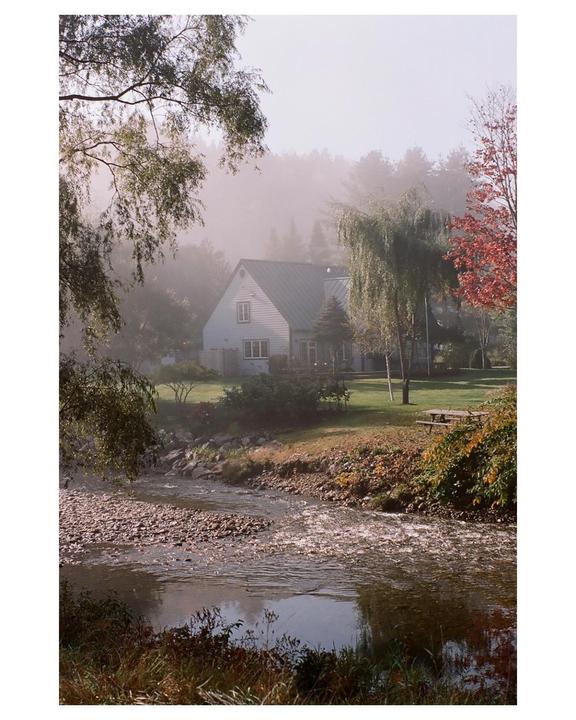 A house is visible through a hazy fog across a rocky stream with autumn foliage.