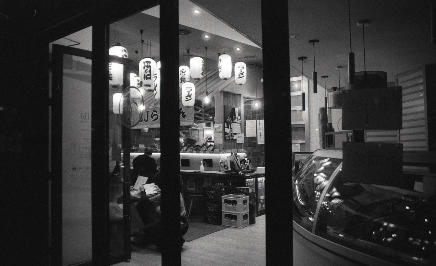 A black and white street photograph shows the interior of a restaurant with hanging lanterns and a man seated at a counter, viewed through a window.