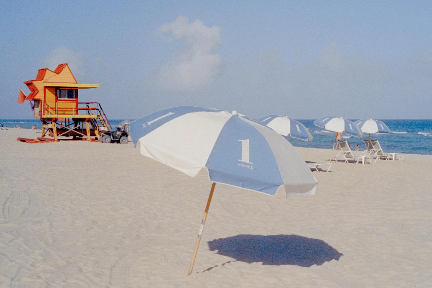 A blue and white beach umbrella casts a shadow on the sand with a lifeguard stand and ocean in the background.