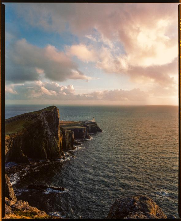 A lighthouse stands on a cliff overlooking the ocean under a cloudy sky at sunset.