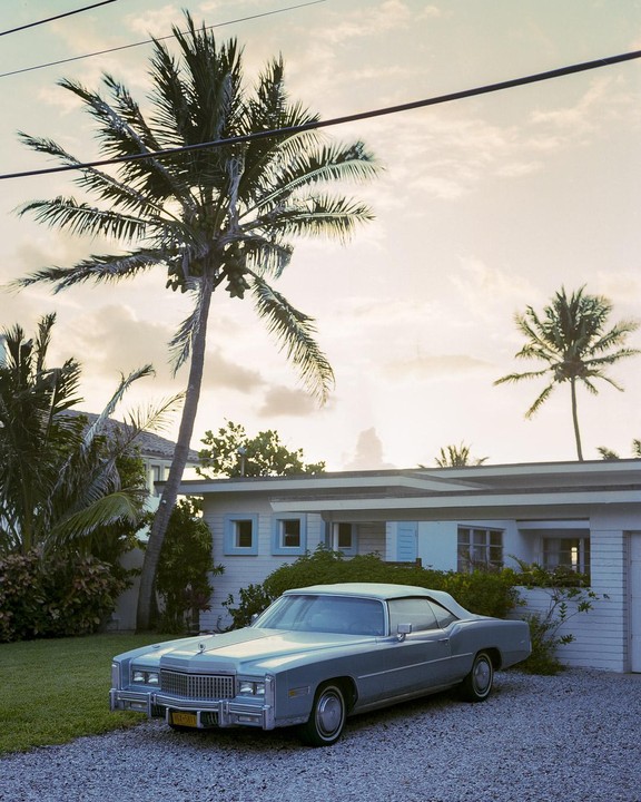 A light blue convertible car is parked on a gravel driveway in front of a white house with palm trees under a cloudy sky.
