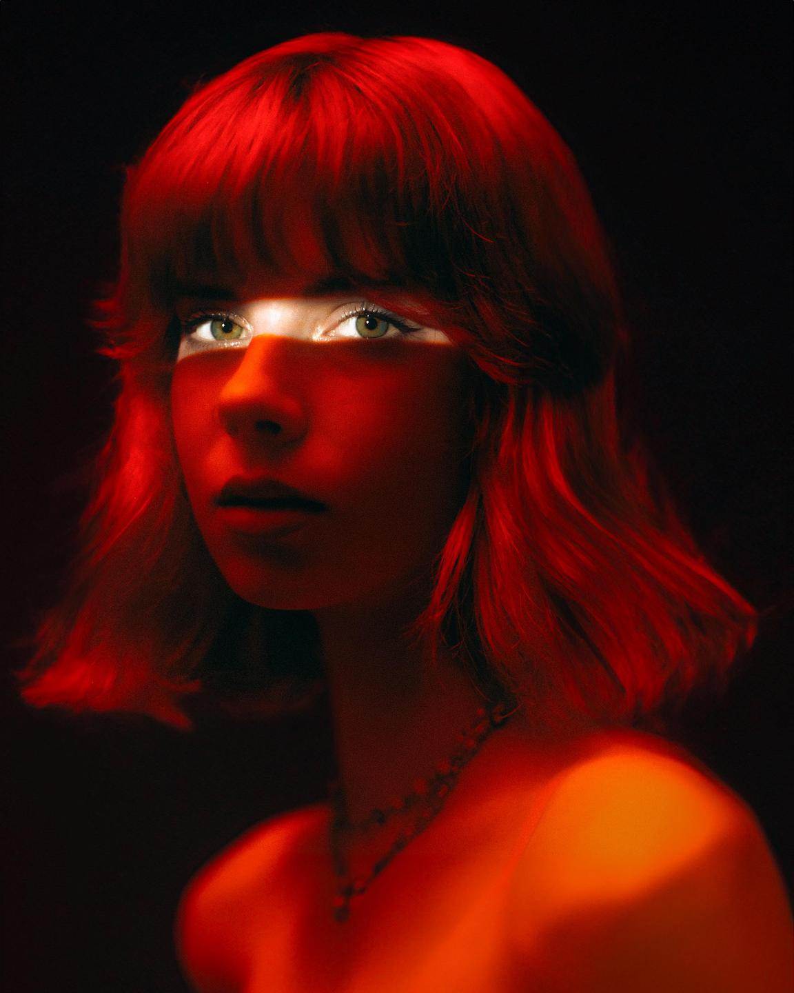 A woman with red hair and a necklace is illuminated by a red light that casts a shadow across her eyes.