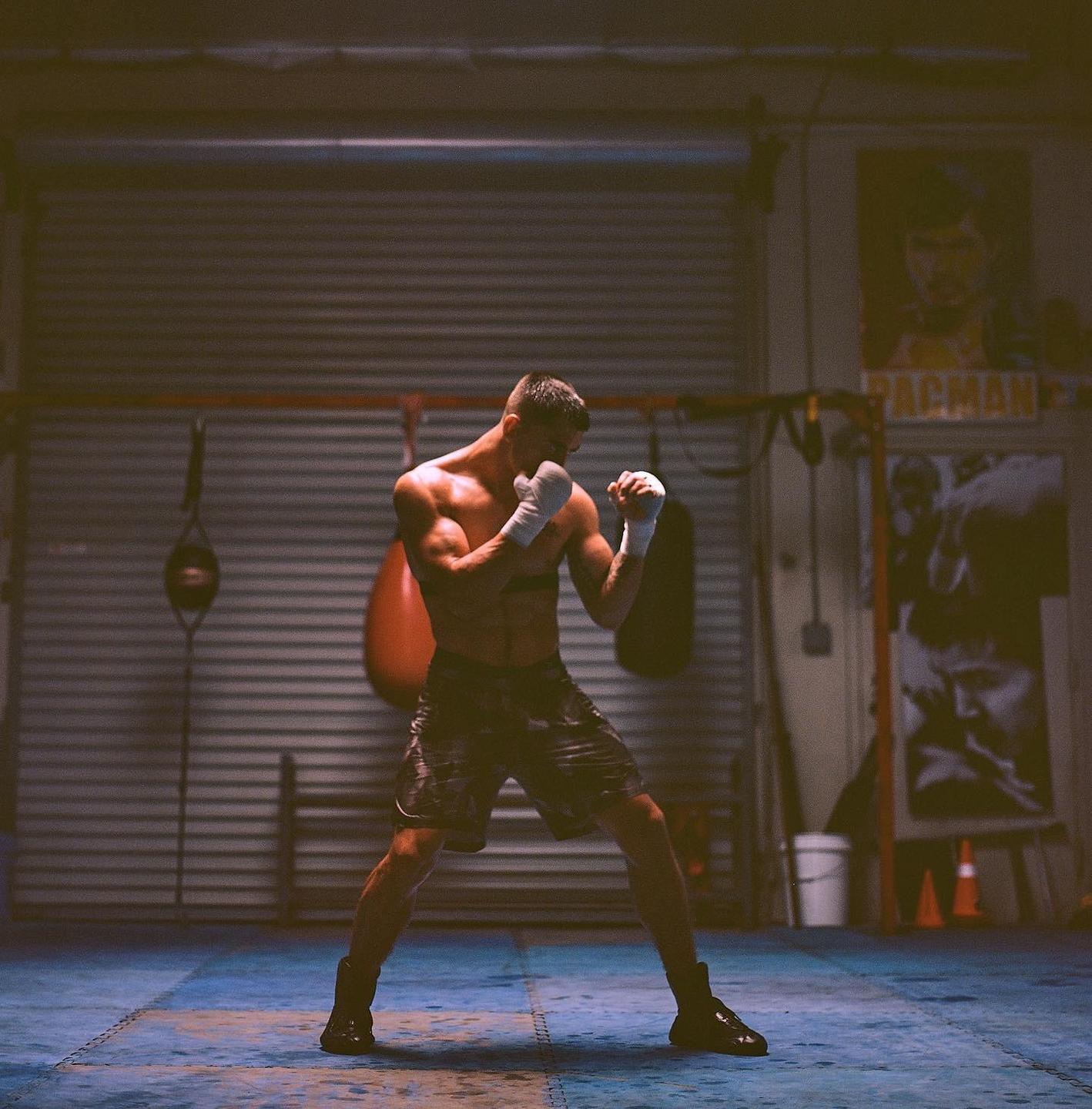 A man in boxing stance is training in a gym with punching bags and a poster in the background.