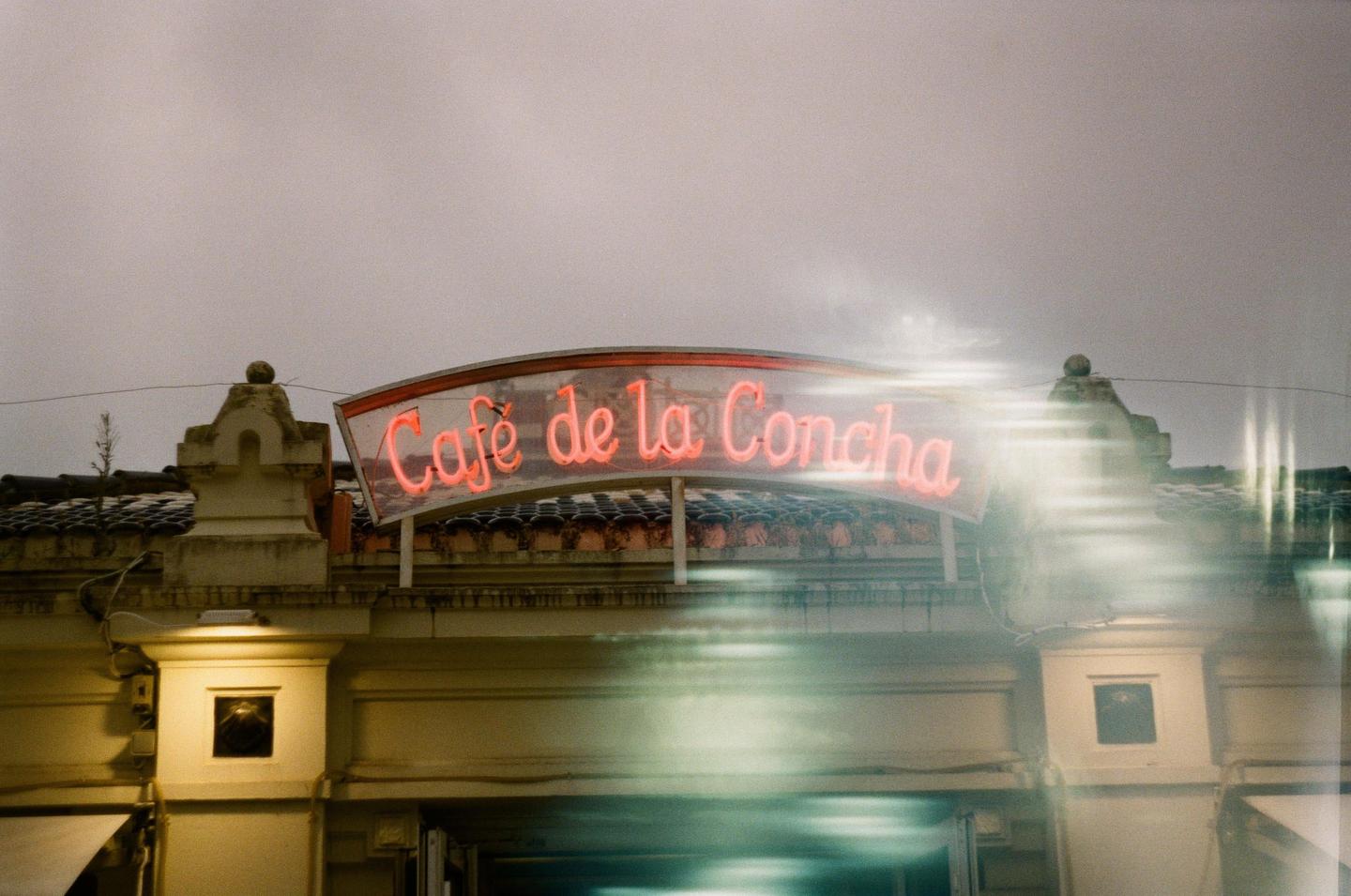 A neon sign for "Cafe de la Concha" is mounted on a building under a cloudy sky.